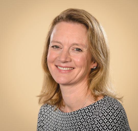 A woman with blonde hair wearing a black and white shirt, smiling and standing against a neutral bac