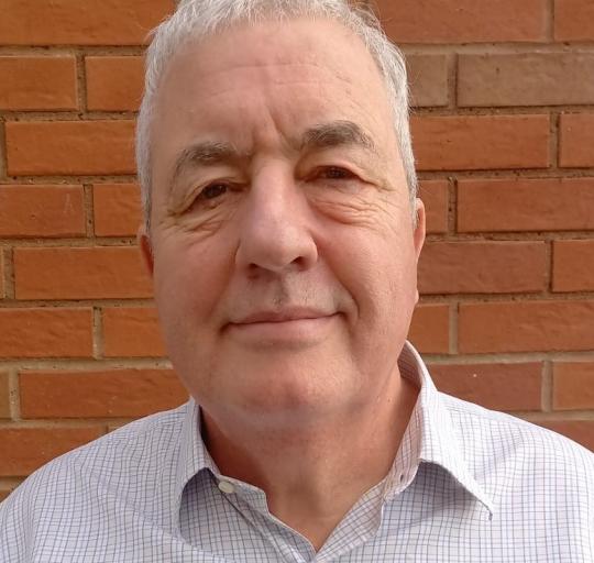  A man wearing a blue and white shirt stands in front of a textured brick wall.