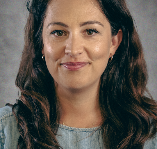 A woman with dark hair smiling against a neutral dark backdrop.