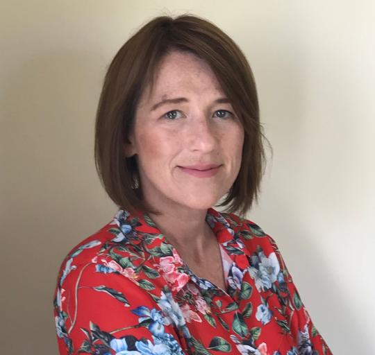 A portrait of a woman with dark mid-length hair wearing a bright, floral top. She is smitling warmly