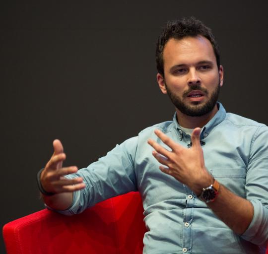  A man seated in a red chair, extending his hands outward in a welcoming gesture.