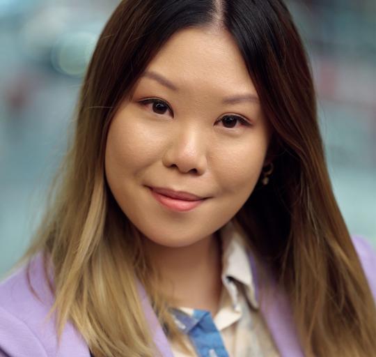 A woman wearing a purple blazer over a white shirt standing in front of a blurred backdrop.