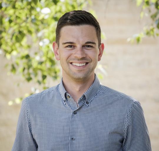 A portrait of a man with short, dark hair, wearing a blue and white checkered shirt. He is smiling w