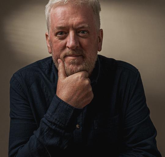A man with a beard dressed in a blue shirt standing in front of a neutral backdrop.