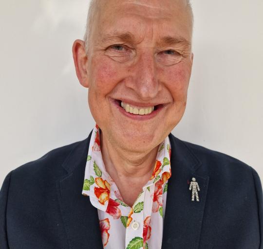 A man with white hair wearing a blue jacket stands against a neutral white backdrop.