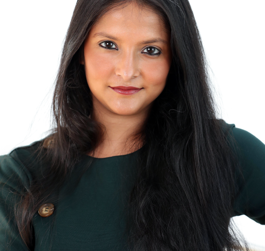 A woman with long hair standing against a light, neutral background.