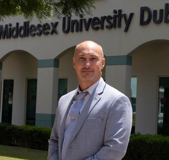  A man with a shaved head wearing a light gray suit and tie stands confidently in front of a buildin