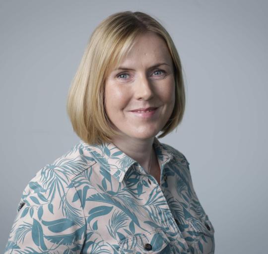 A woman with short blonde hair wearing a vibrant floral shirt, smiling against a neutral background.