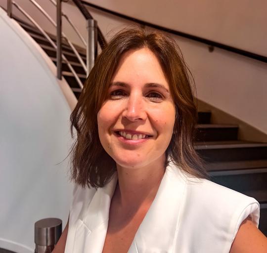 A woman wearing a white blazer stands in front of a stairway.