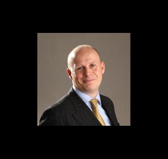 A bald man dressed in a formal suit and tie against a neutral background.