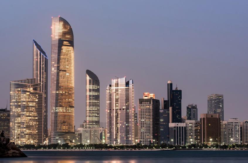 A city skyline at night with tall skyscrapers illuminated against a dark sky, reflecting on the water of a nearby lake or river. The scene captures the essence of a bustling metropolis with bright lights and urban architecture.