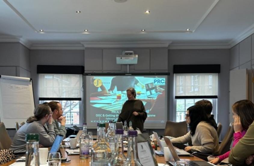 A female trainer with blond hair in black clothing standing in front of a projector screen in a training session